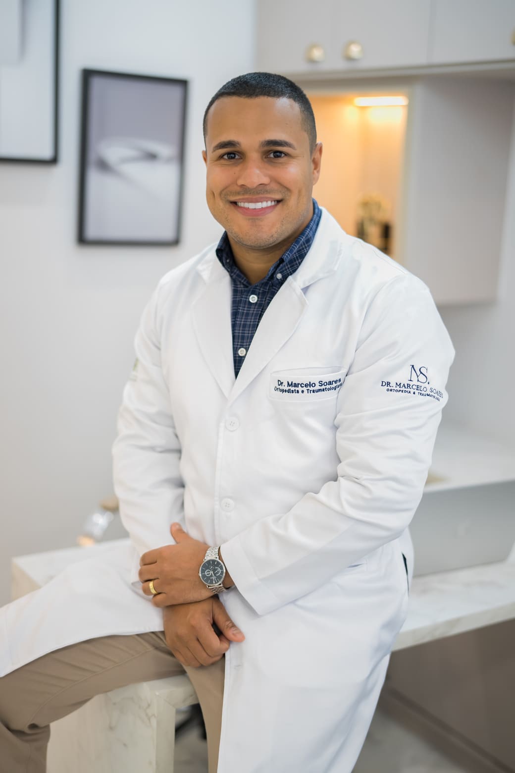 Dr. Marcelo Soares, ortopedista, sorrindo de jaleco branco bordado, sentado em consultório moderno com quadros e iluminação suave, fotografia profissional ultra-realista