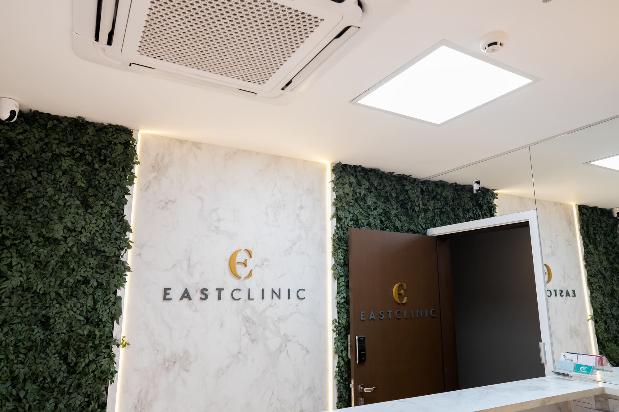 Recepção da EastClinic com parede de mármore iluminada, painel verde de folhagem e logotipo dourado, ambiente clean e sofisticado, fotografia de interiores ultra-realista