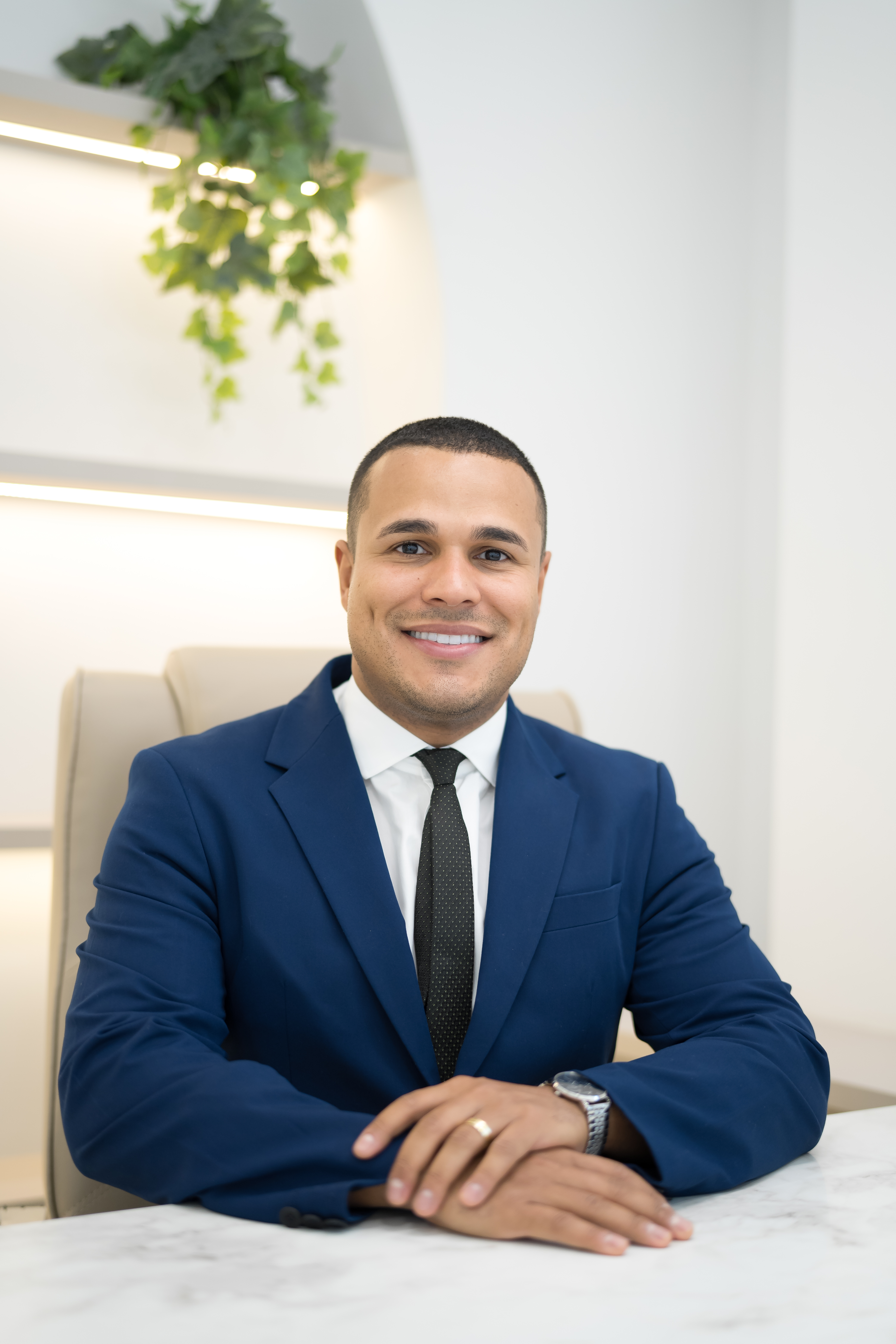 Dr. Marcelo Soares, ortopedista, sorrindo de terno azul-marinho, mãos entrelaçadas sobre mesa de mármore branco com planta pendente ao fundo, fotografia profissional ultra-realista